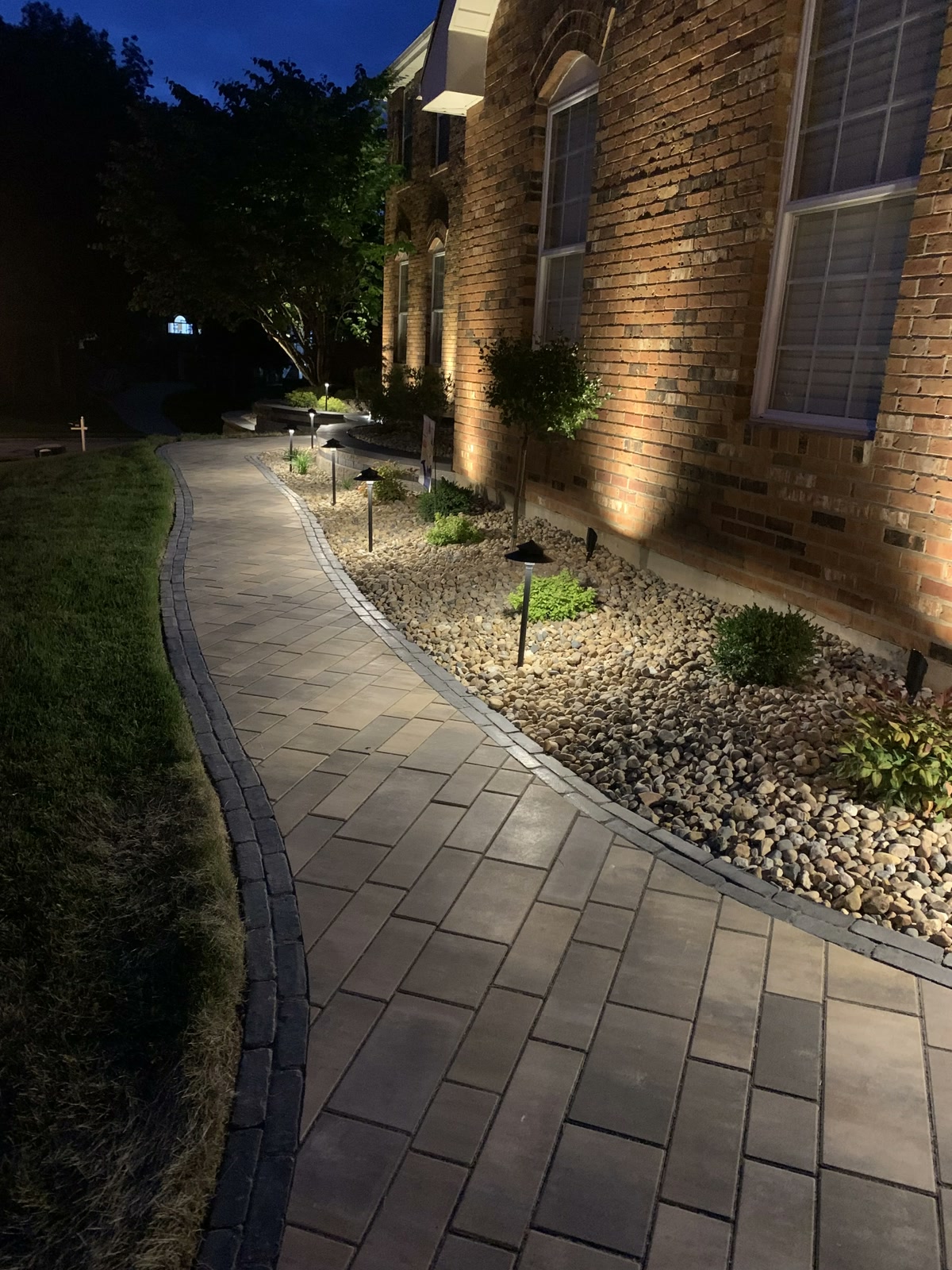 Upright view of a curved paver walkway with path lighting and home uplighting at dusk